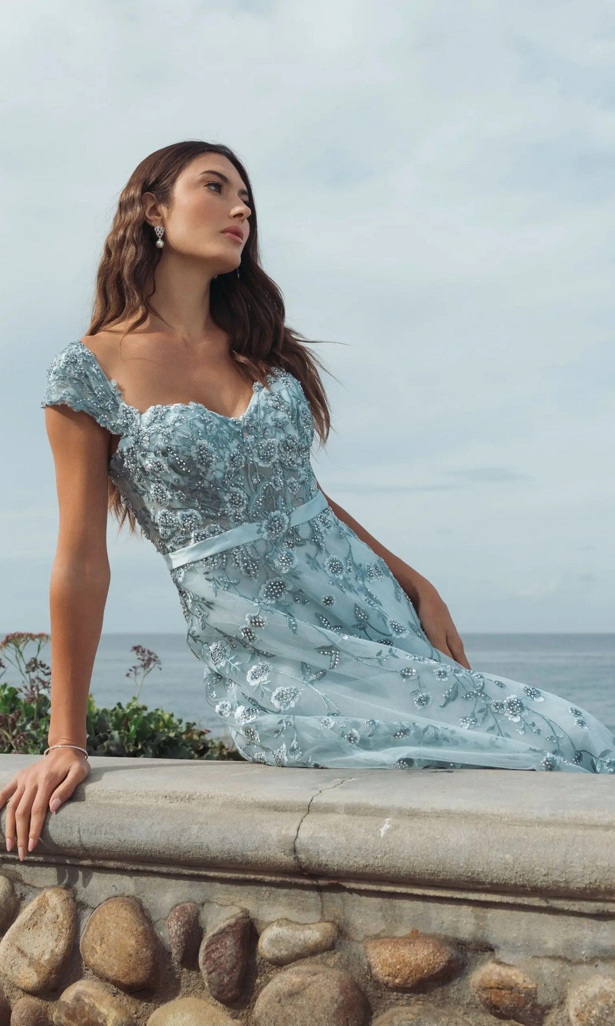 Woman in a Cap-Sleeve Nude Midi Party Dress (Mac Duggal 20582) leans against a stone ledge outdoors, with the ocean and cloudy sky in the background.